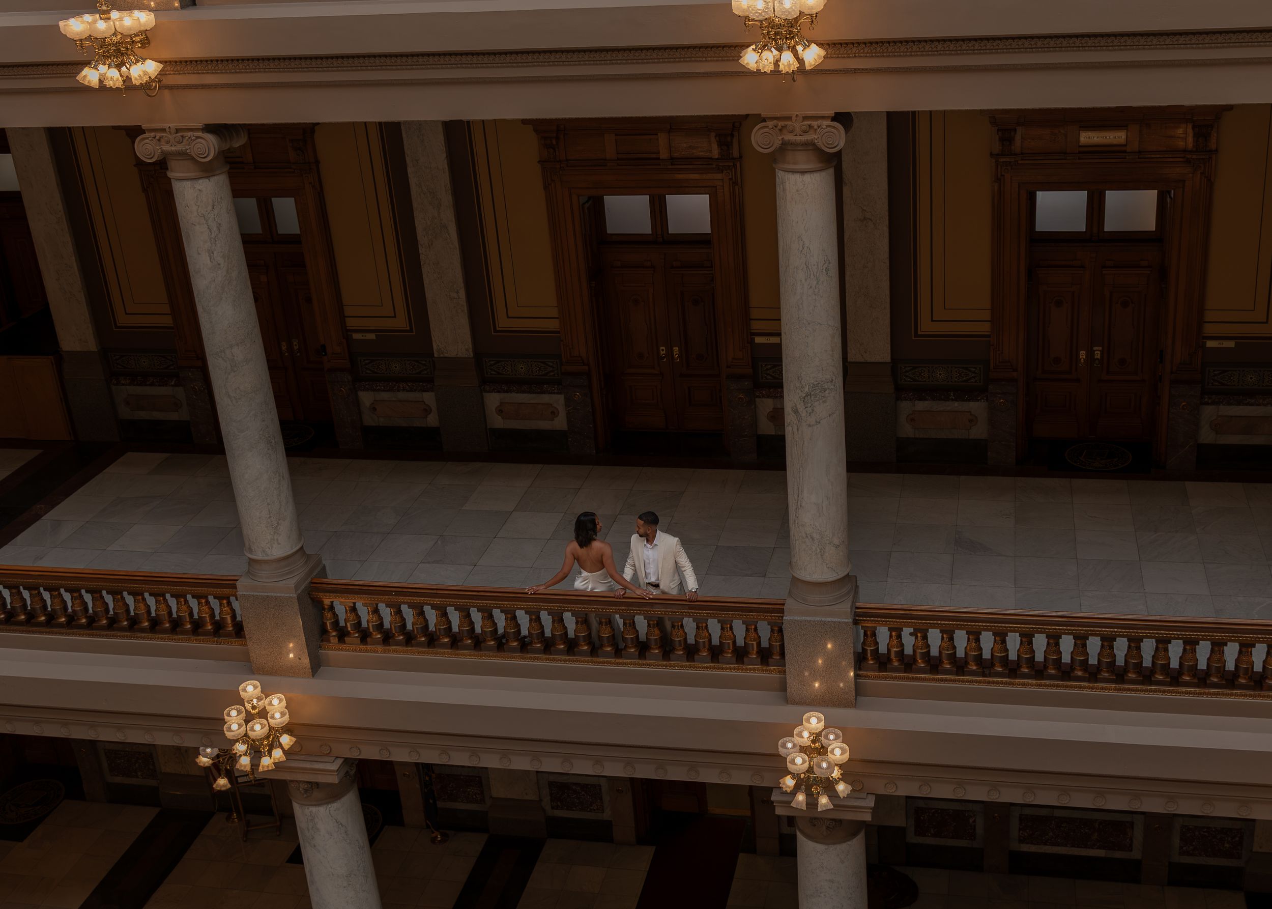 Couple sharing a romantic moment on a grand balcony with marble columns and chandeliers in an elegant historic building.
