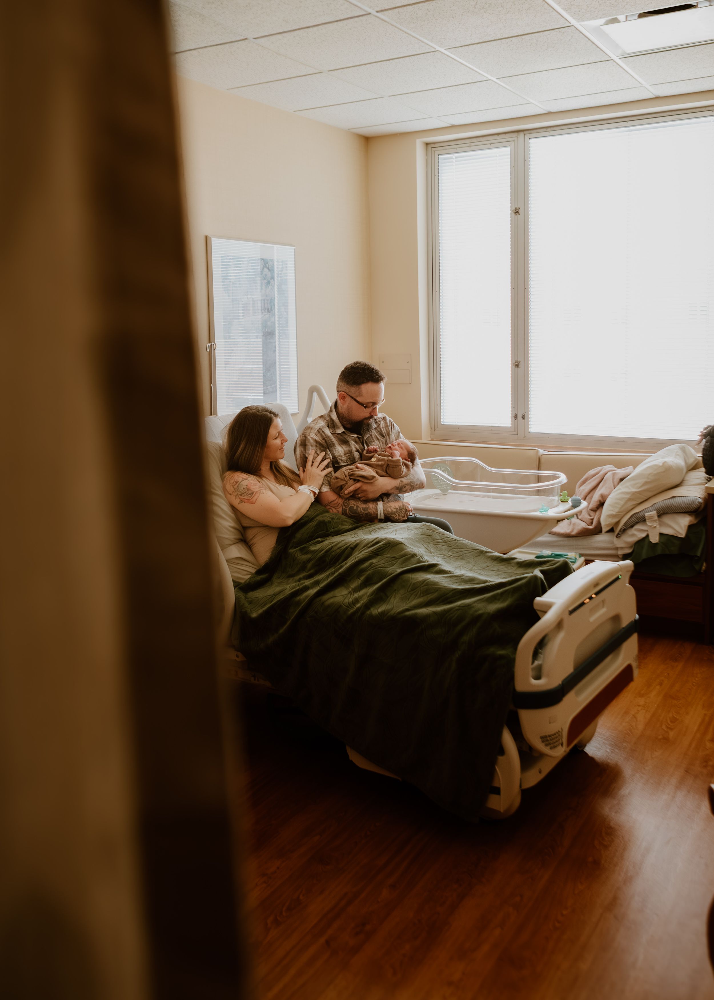 New parents sitting together in hospital bed lovingly holding their newborn baby by a bright window.