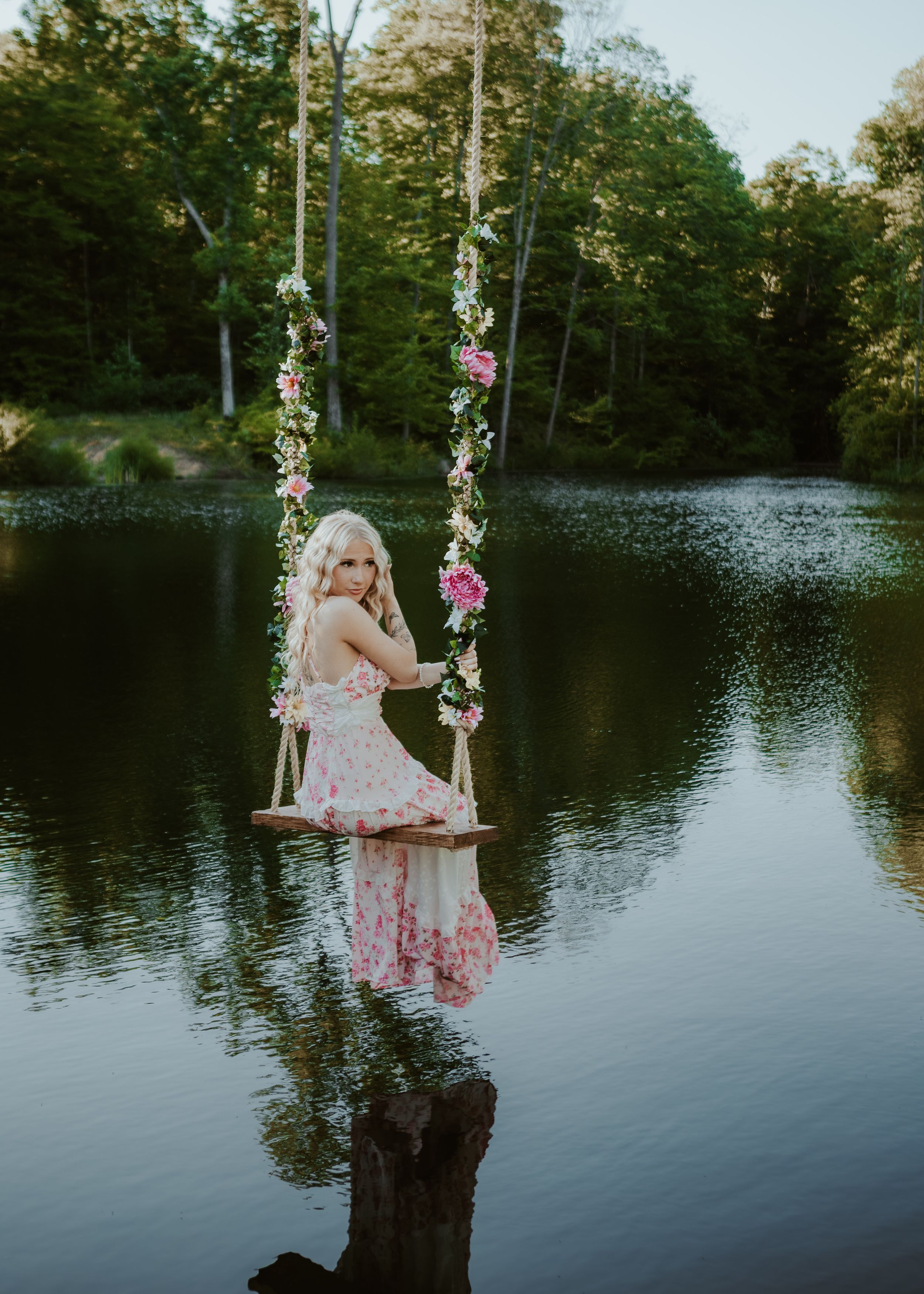 Blonde woman in floral pink dress sitting on flower-adorned swing suspended over a serene forest lake.