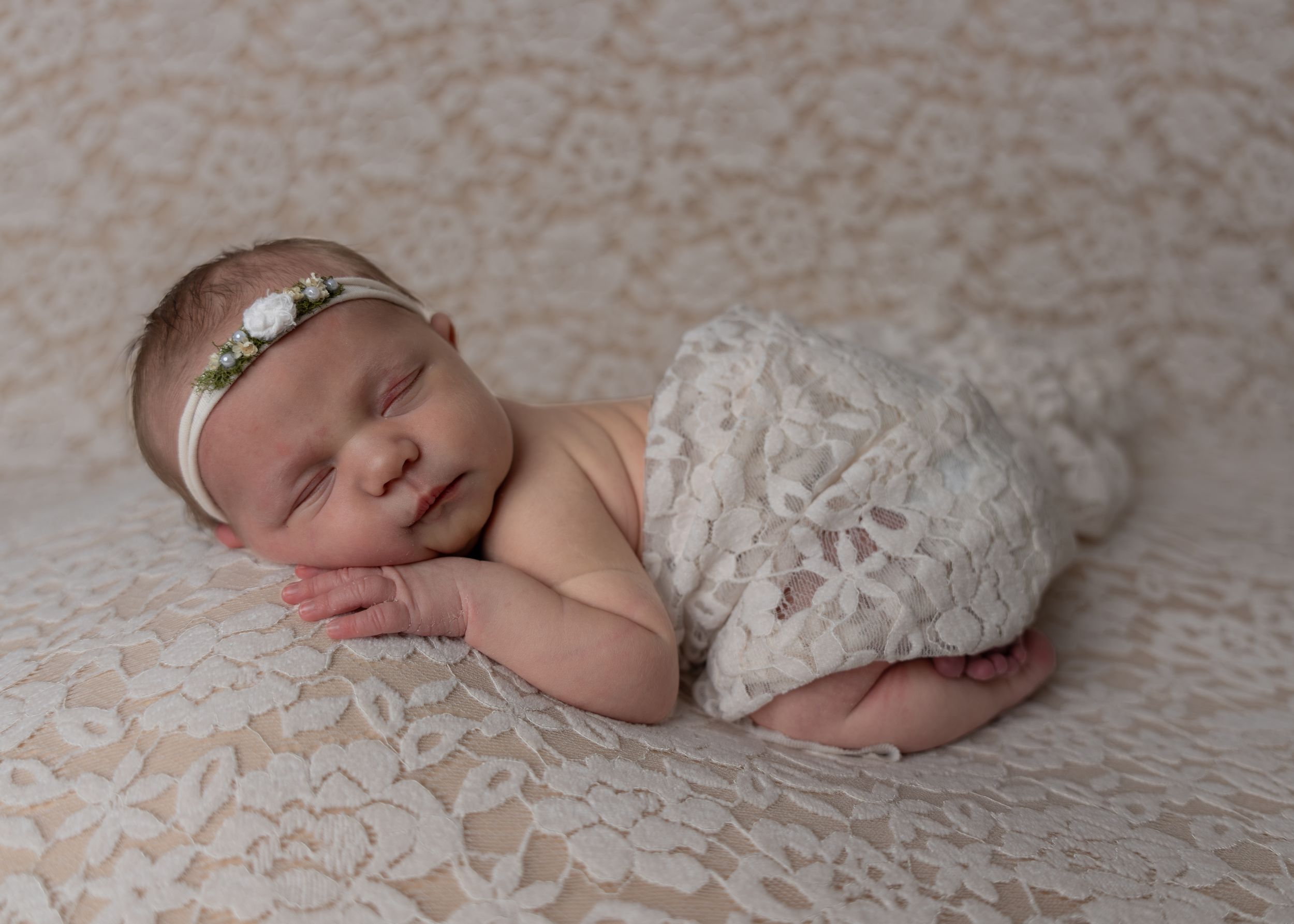 Peaceful newborn baby girl sleeping in lace wrap with floral headband on matching lace backdrop.