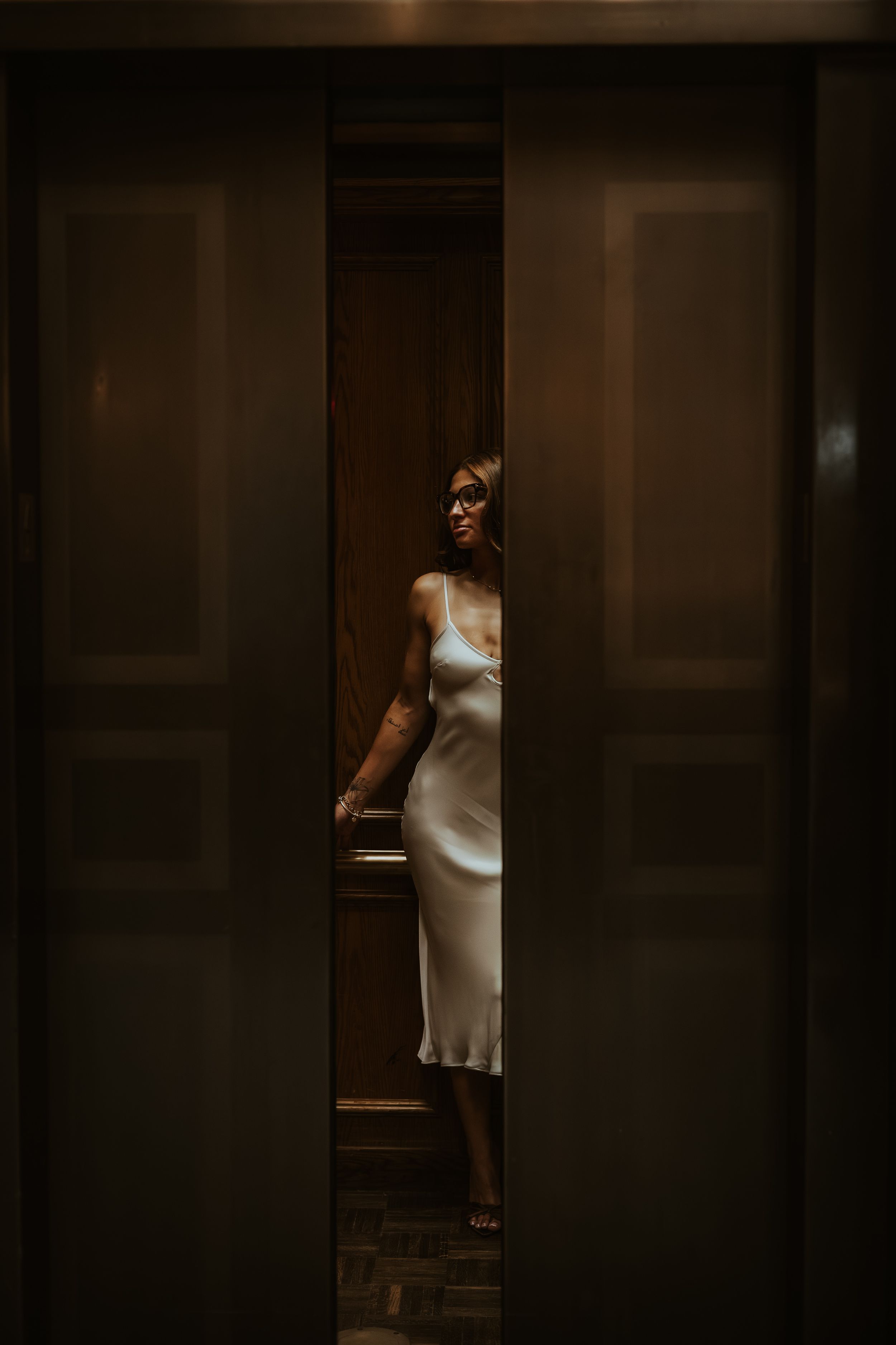 Elegant woman in white satin slip dress standing in dimly lit doorway of a sophisticated interior.