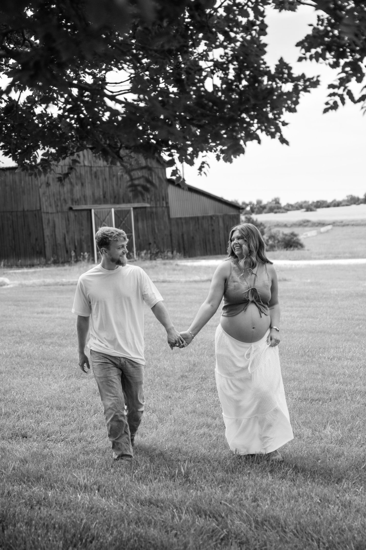 Expecting couple holding hands walking through green field near rustic barn in black and white maternity photo.