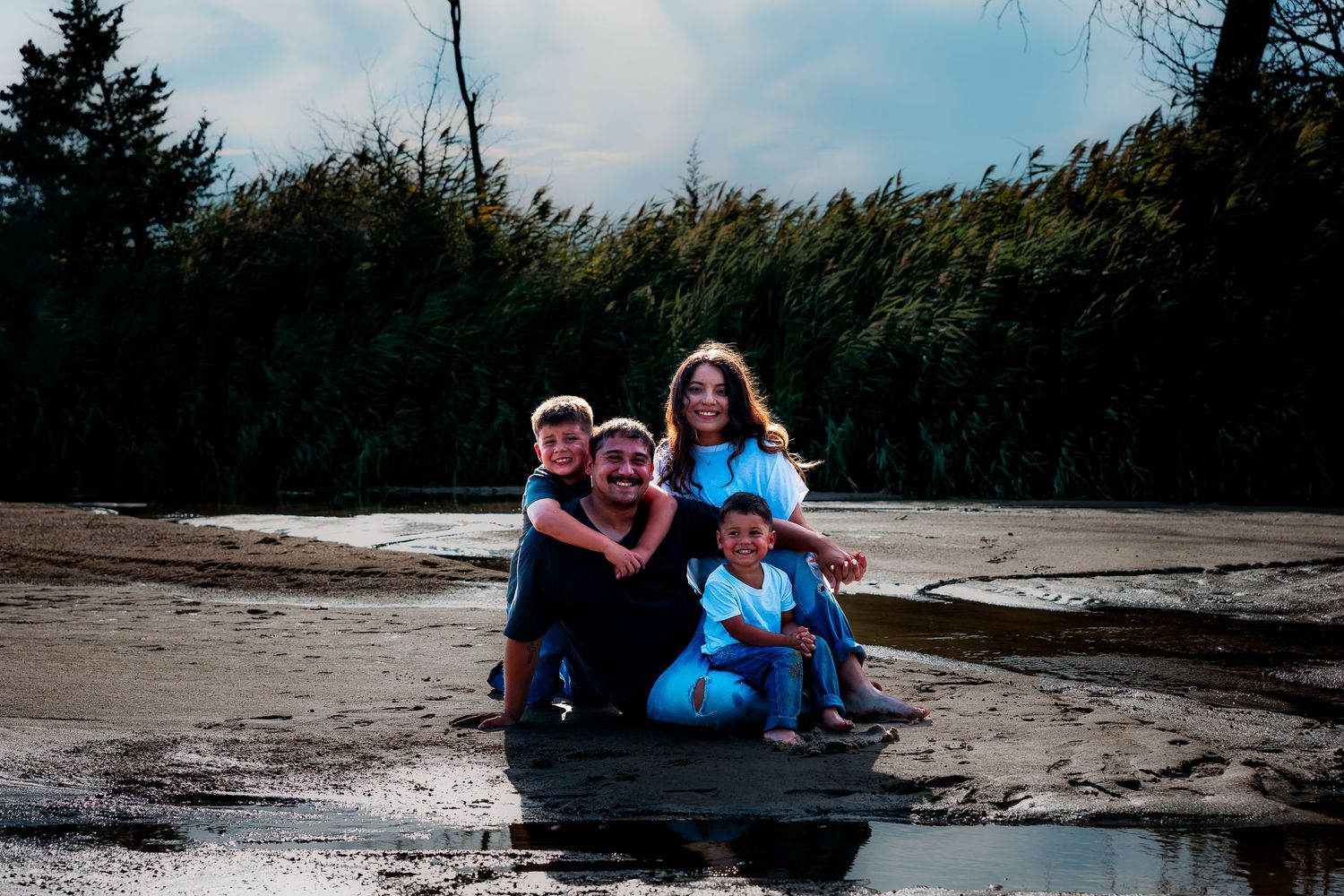 Family of four standing barefoot in shallow water at a sandy beach, parents smiling at each other while children stand beside them.