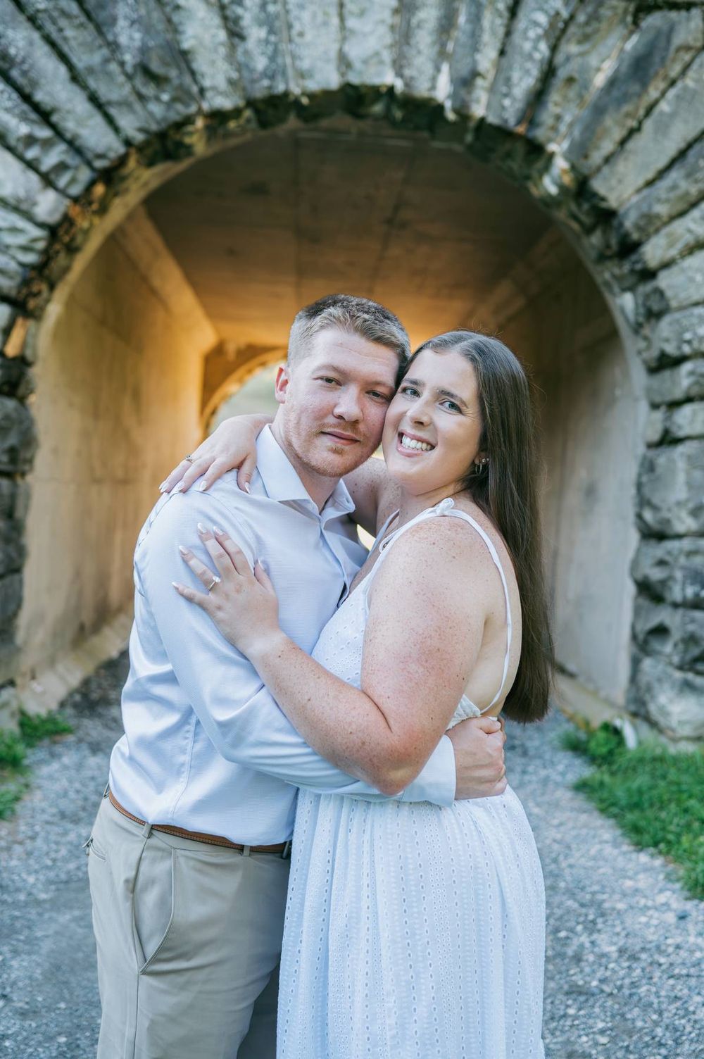 Smiling engaged couple embracing under glowing stone arch tunnel, woman in white dress showing engagement ring, engagement photographer NC