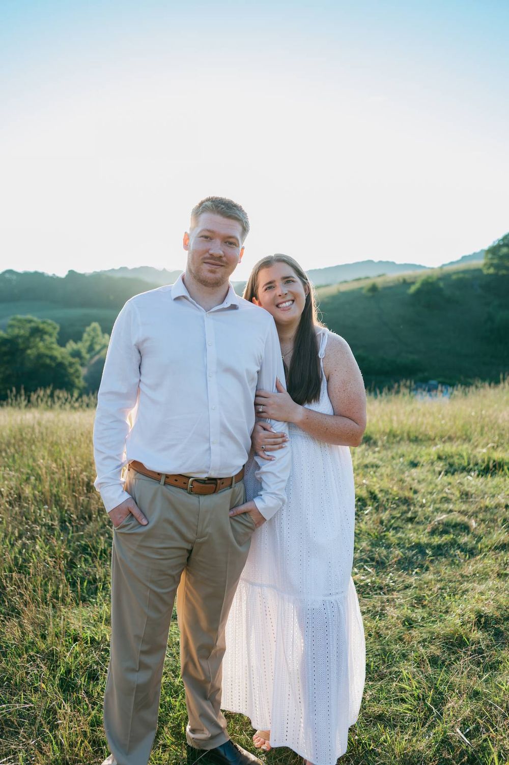 Happy couple standing in golden sunlit meadow with rolling hills, woman in white dress holding mans arm, engagement photographer NC