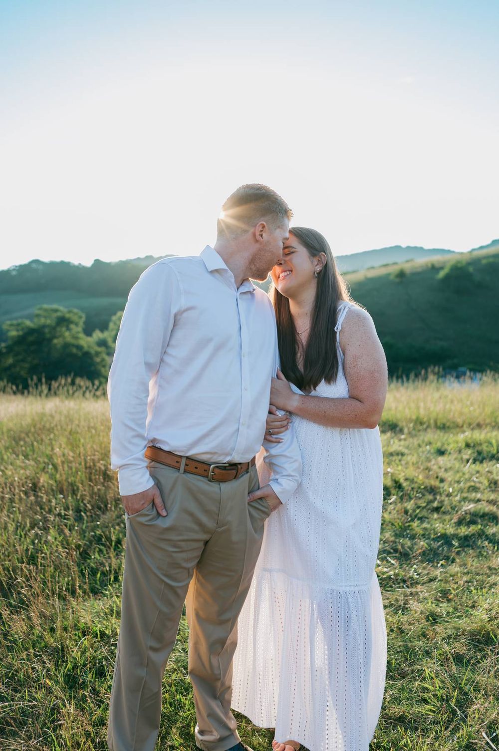 Couple sharing a gentle kiss in a sunlit grassy field with green hills and bright sky behind them, engagement photographer North Carolina