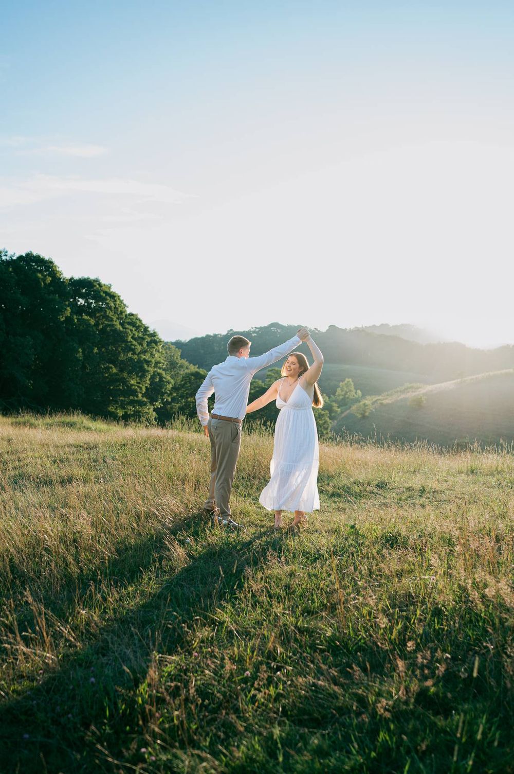 Couple dancing in golden sunlit meadow, man spinning woman in flowing white dress with scenic hills backdrop, engagement photographer NC