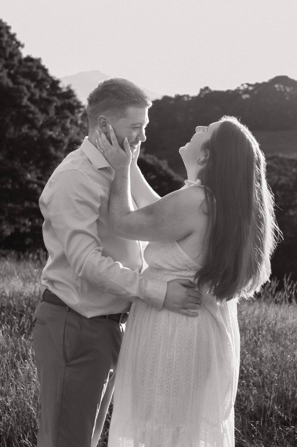 Black and white photo of a couple laughing and embracing in a field at golden hour, engagement photographer North Carolina
