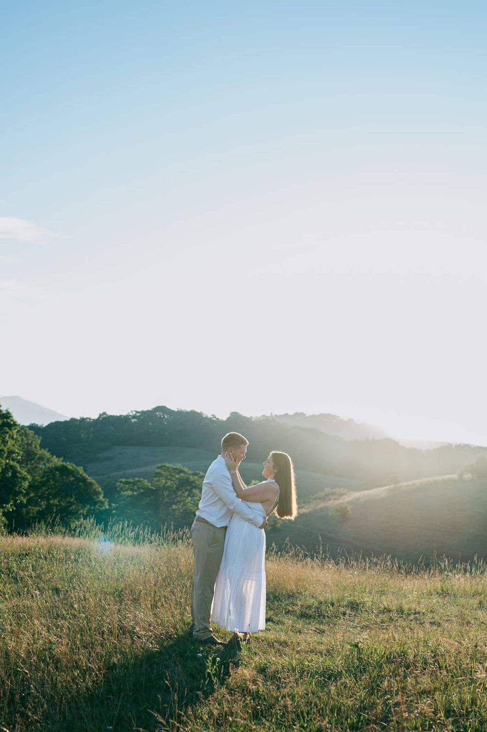 Couple embracing in a sunlit meadow with rolling hills and golden sunset behind them, engagement photographer High Point NC