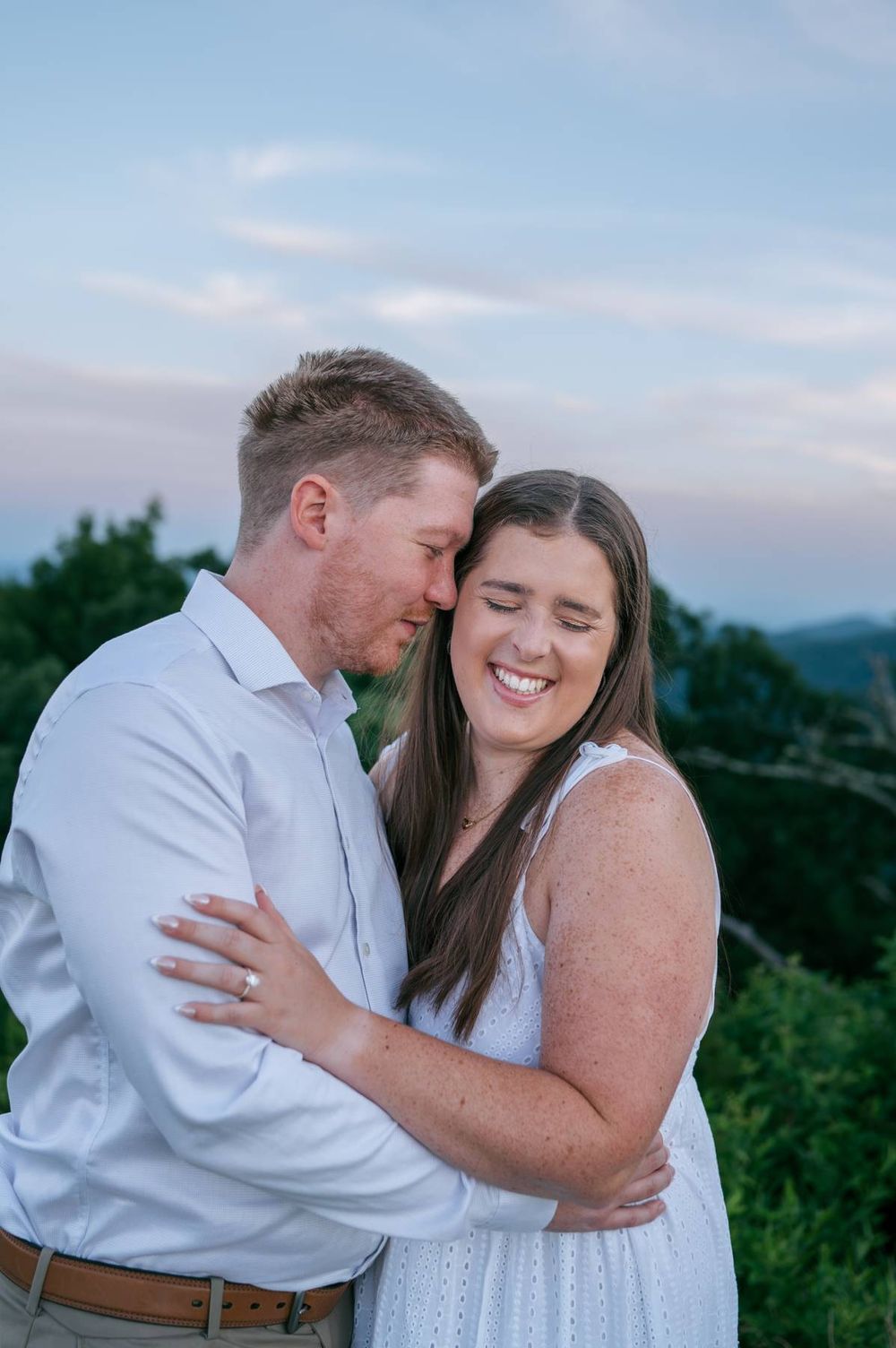 Smiling couple cuddling at sunset with lush green mountains in the background, engagement photographer Asheboro NC