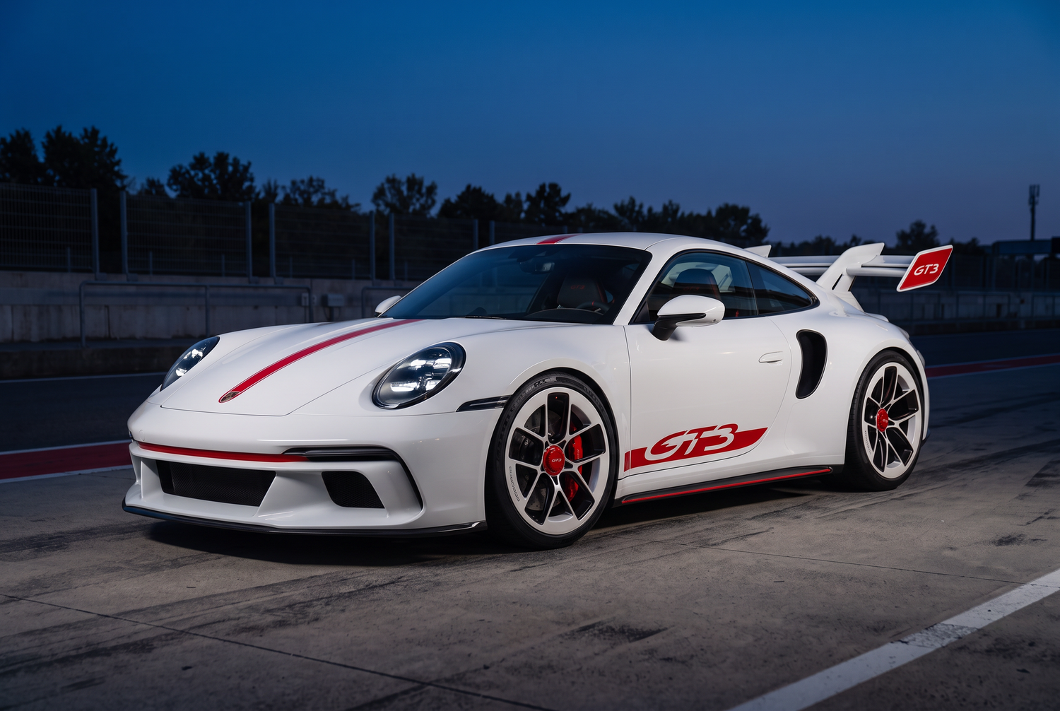 White Porsche 911 GT3 RS sports car with red accents parked on a race track at dusk.
