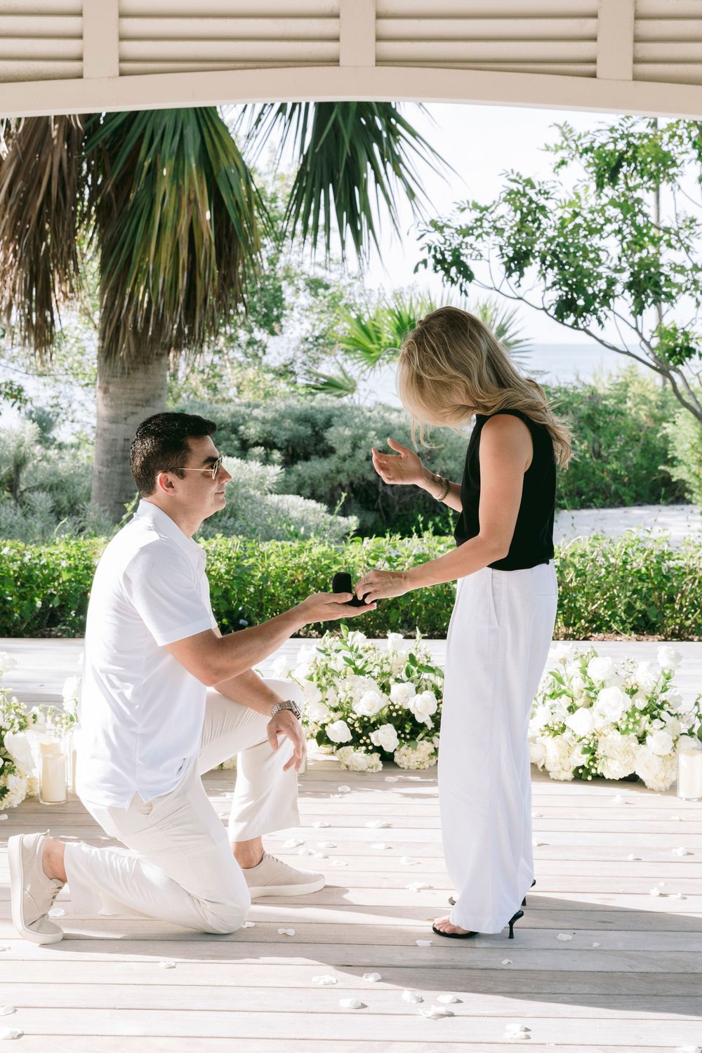 Man kneeling on one knee proposing to woman with ring box on elegant outdoor terrace surrounded by white flowers and palm trees.