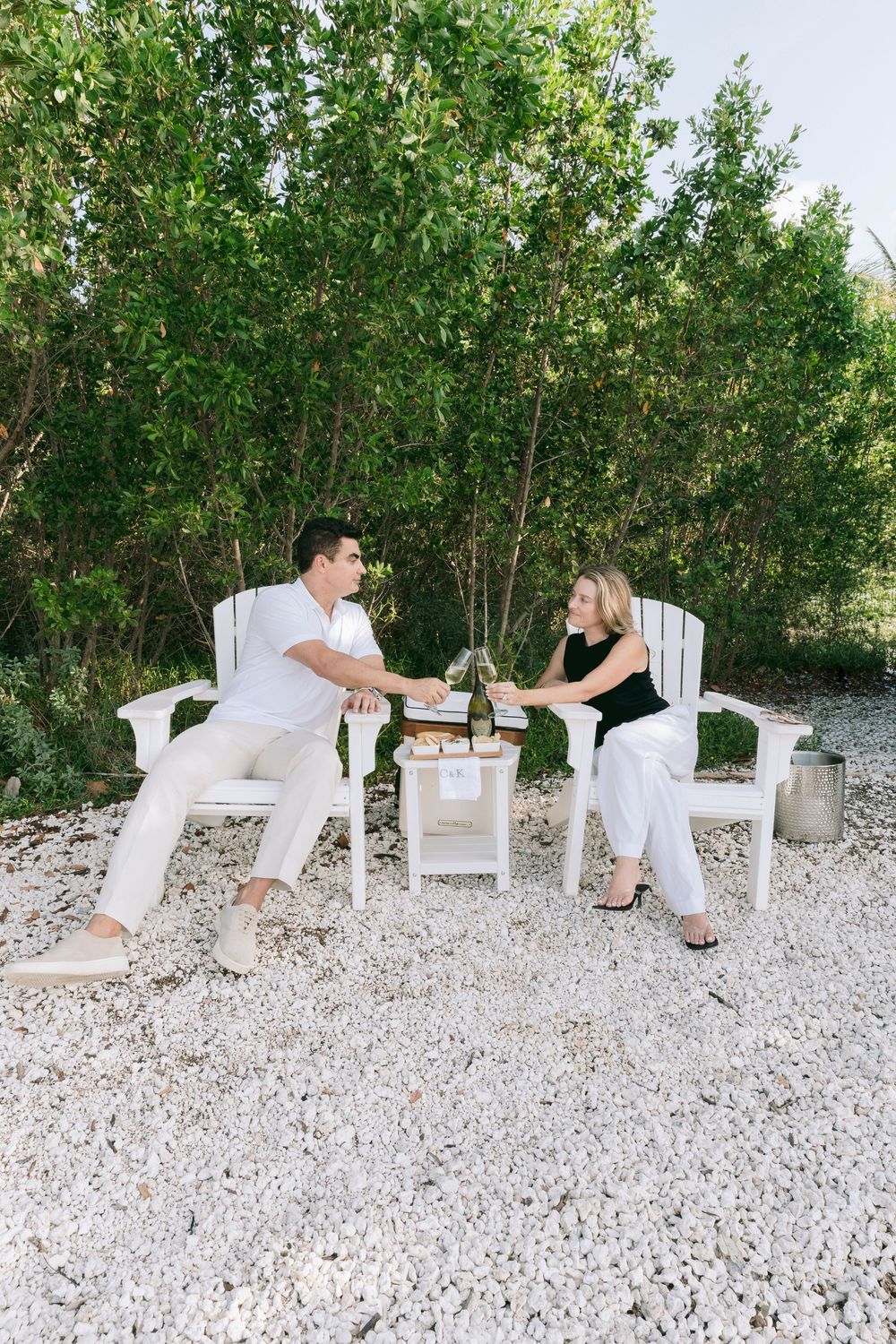 Couple toasting with wine glasses on white Adirondack chairs surrounded by gravel and lush green trees outdoors.