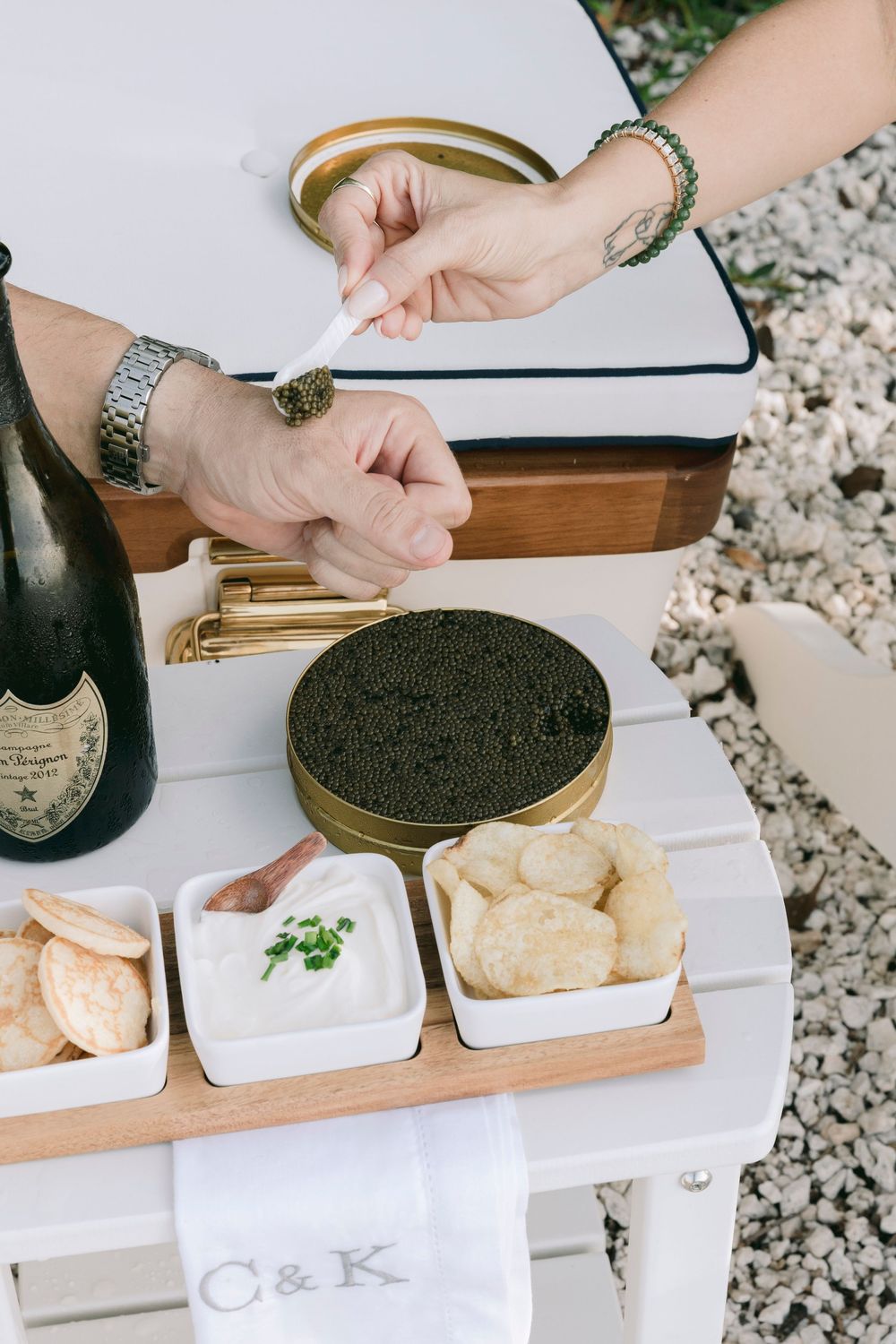 Hands serving black caviar from a tin on a white table with crackers, blinis, cream, and Dom Perignon champagne.