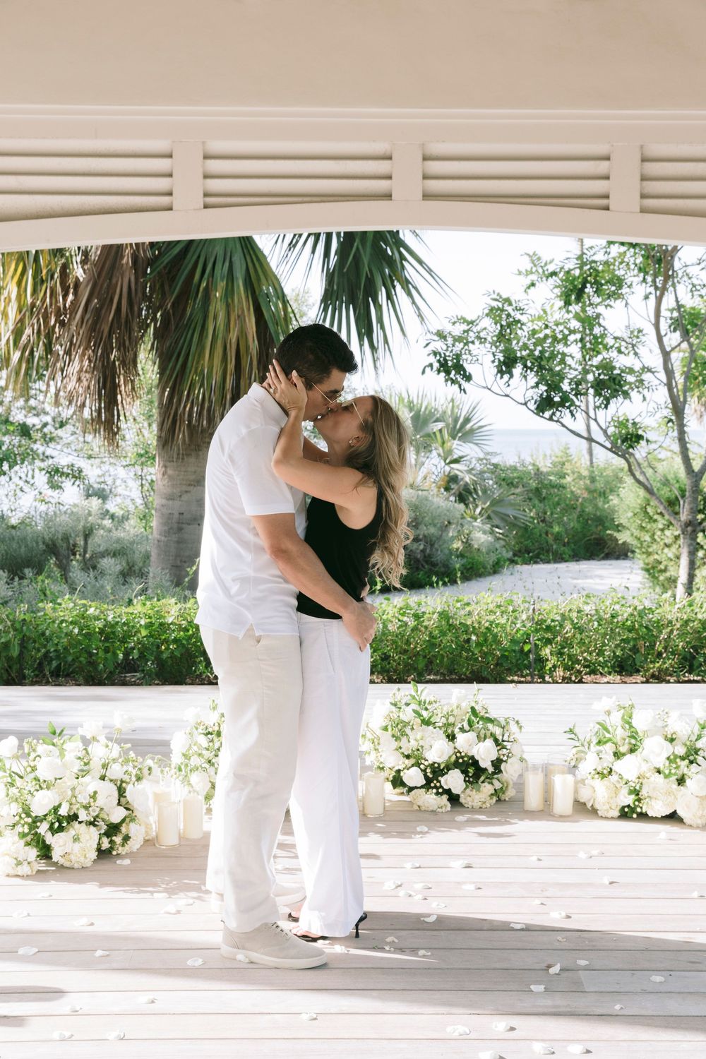 Couple sharing a romantic kiss under a white gazebo surrounded by white floral arrangements and candles outdoors.