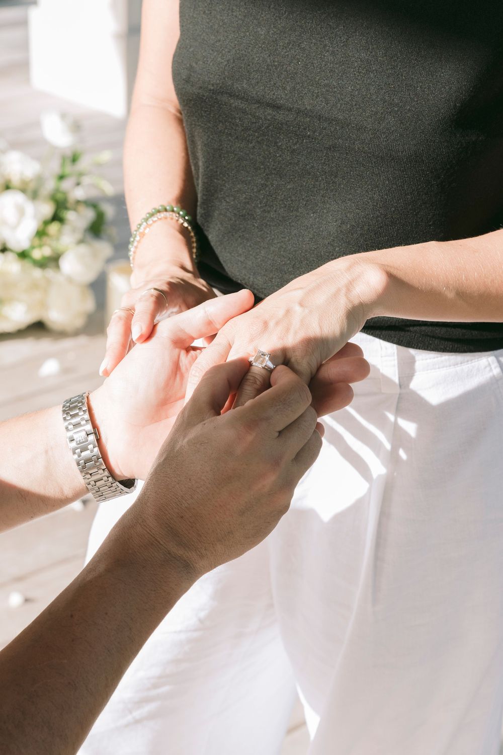 A person placing a diamond engagement ring on their partner's finger during an outdoor wedding ceremony in sunlight.