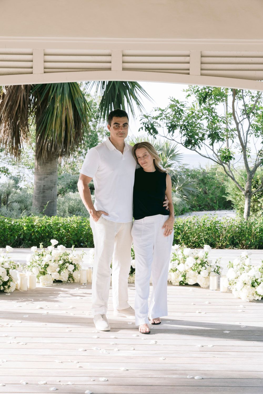 A couple poses under a white pergola surrounded by lush greenery and white floral arrangements on a sunny day.