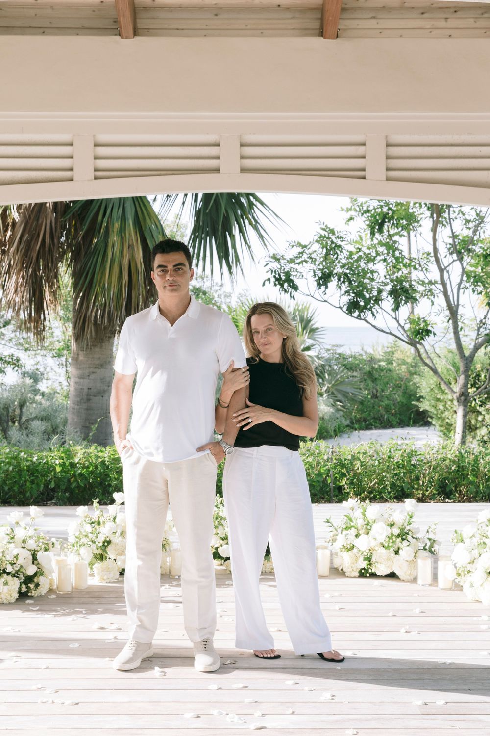 Couple posing under a white pavilion surrounded by lush greenery and white floral arrangements outdoors.