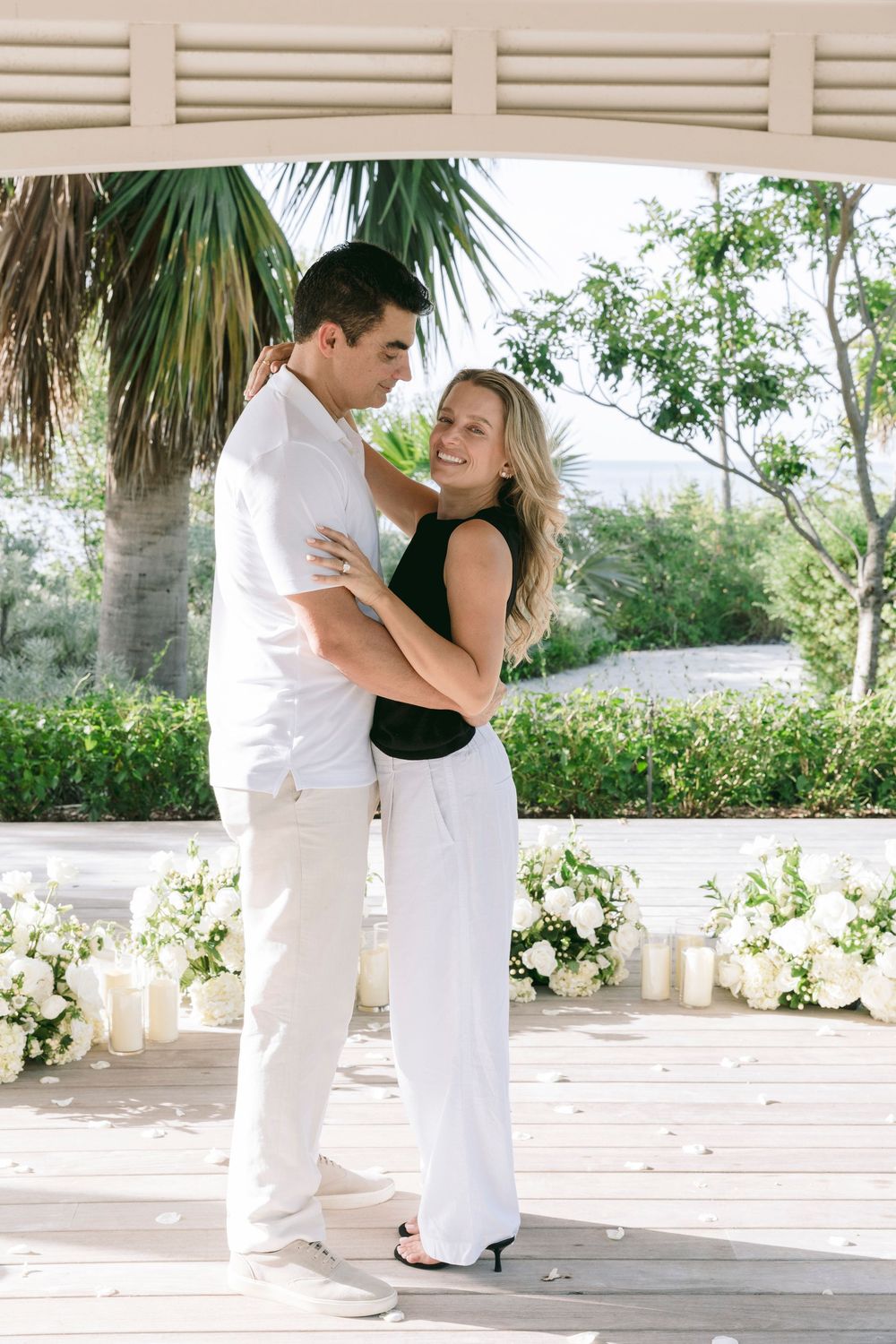 Couple embracing under a gazebo surrounded by white floral arrangements and candles at an outdoor venue.