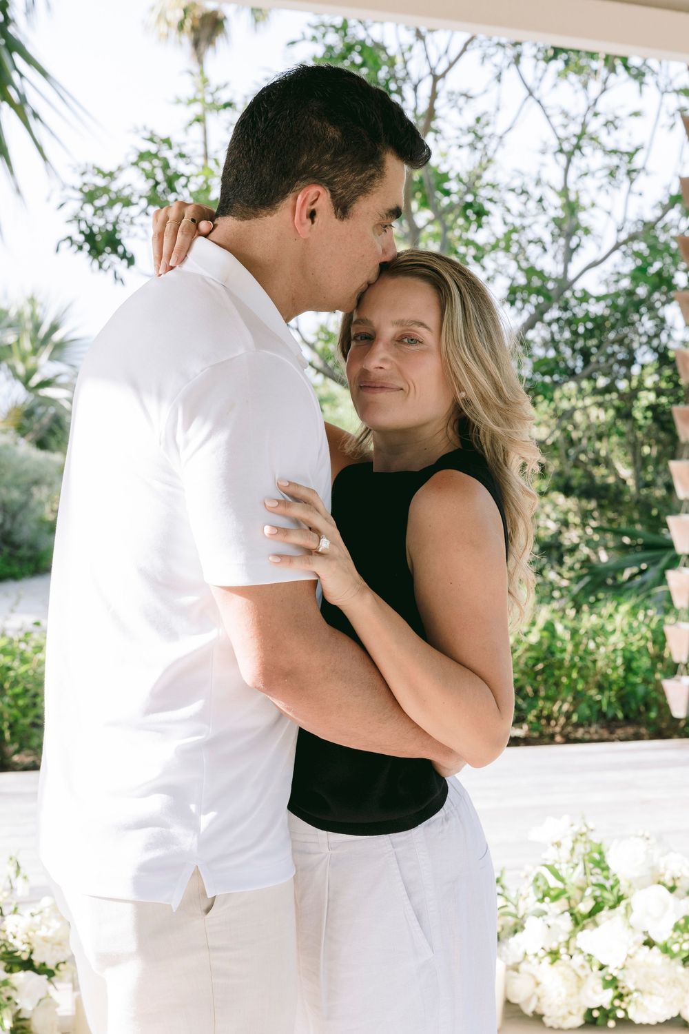 A couple embraces tenderly outdoors, woman in black top and white pants smiling, man in white shirt, surrounded by lush greenery.