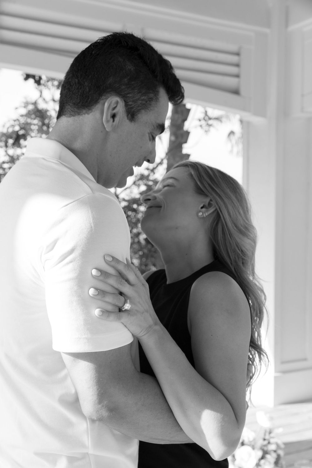 Black and white photo of a romantic couple embracing and laughing together on a porch, woman wearing engagement ring.