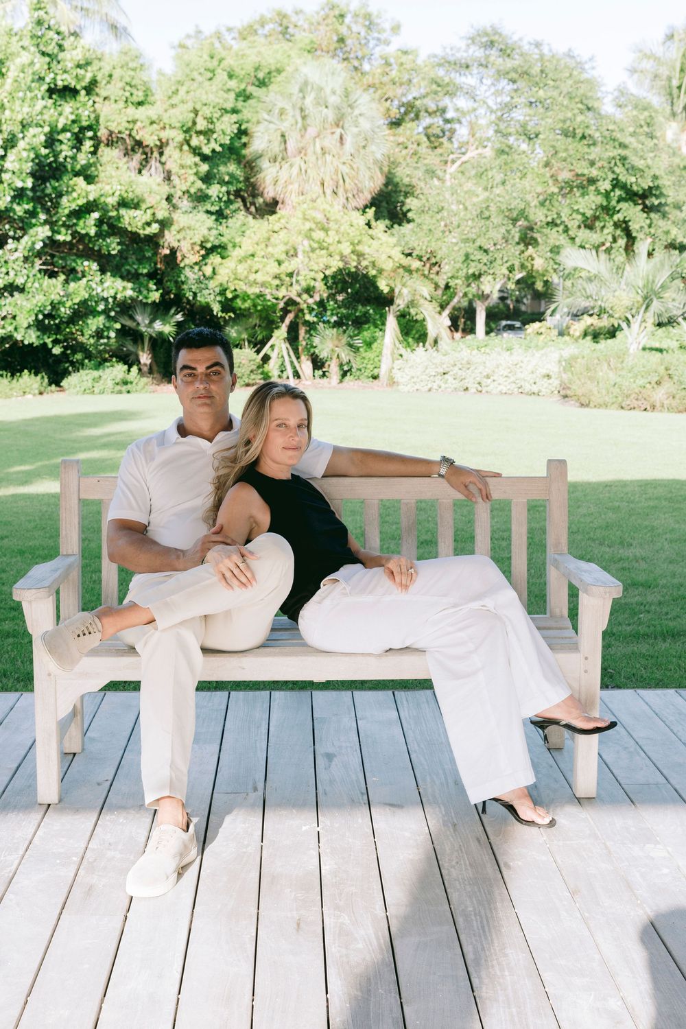 Couple sitting on a white wooden bench outdoors, dressed in white and black outfits, surrounded by lush green trees.