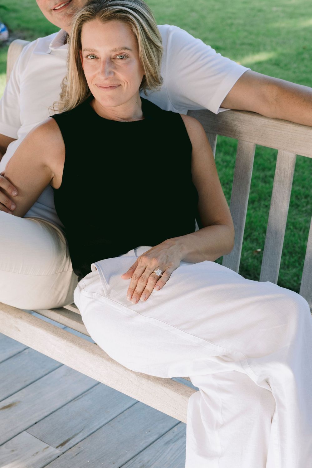 Woman in black top and white pants sits on wooden bench outdoors, leaning against a companion on a sunny day.