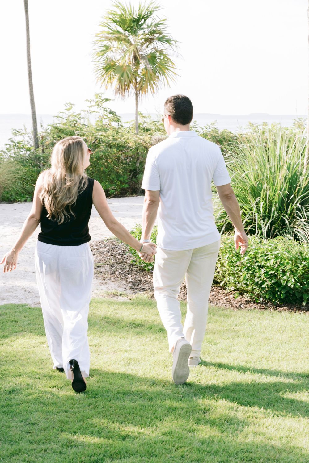 Couple holding hands walking on green lawn toward palm tree and ocean view, woman in black top and white pants.