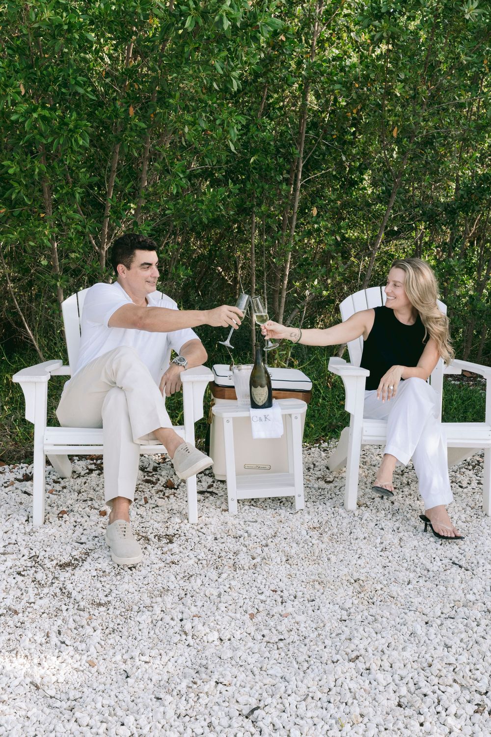 Couple toasting champagne glasses in white Adirondack chairs on a gravel patio surrounded by lush greenery.