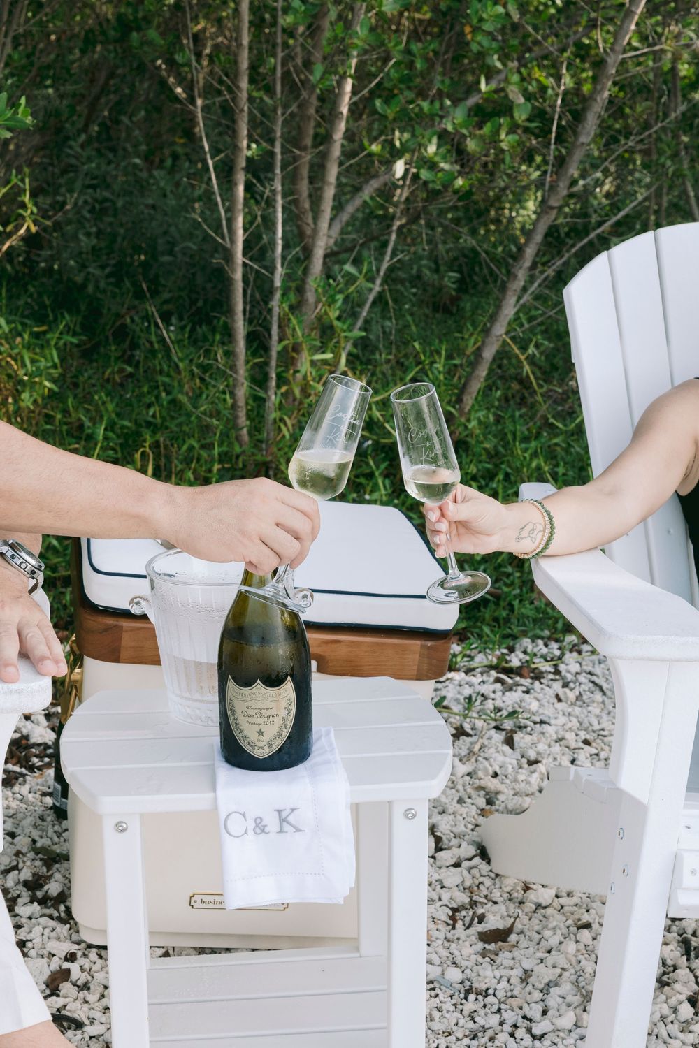 Two people toasting with Dom Perignon champagne flutes on white Adirondack chairs with monogrammed C&K towel.