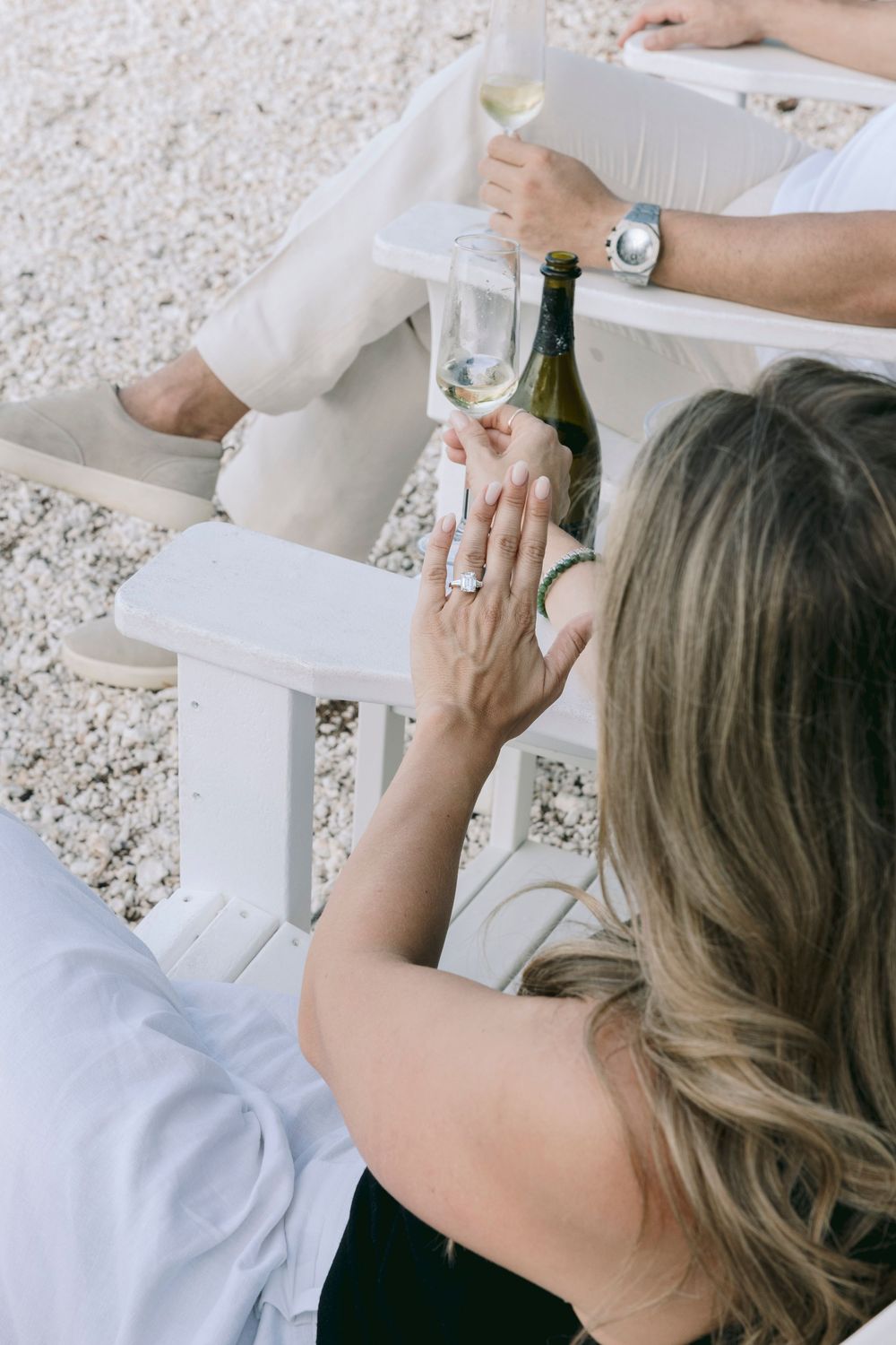 Two people relaxing outdoors with white wine glasses and a bottle, woman wearing a diamond ring, seated in white chairs.