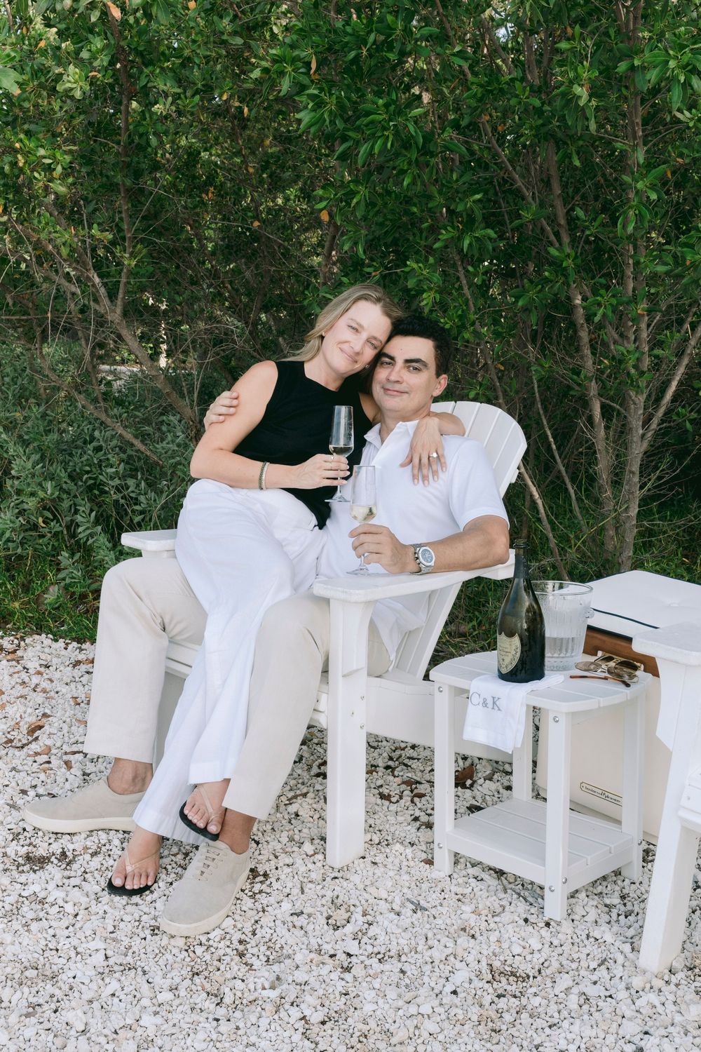Couple sitting together on white chair outdoors, holding champagne glasses, with bottle on side table.