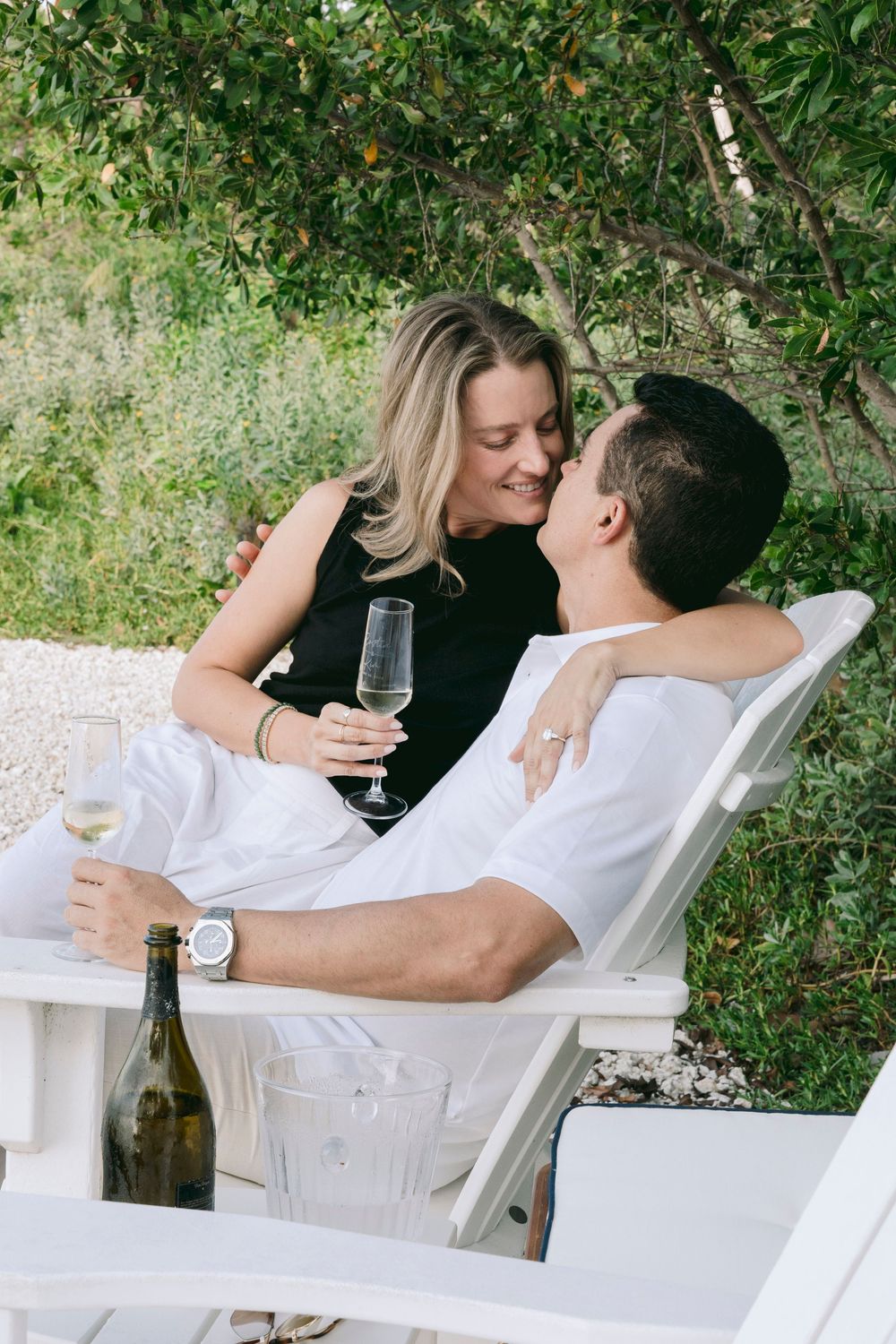 Romantic couple sharing champagne on white lounge chairs in a lush garden setting.