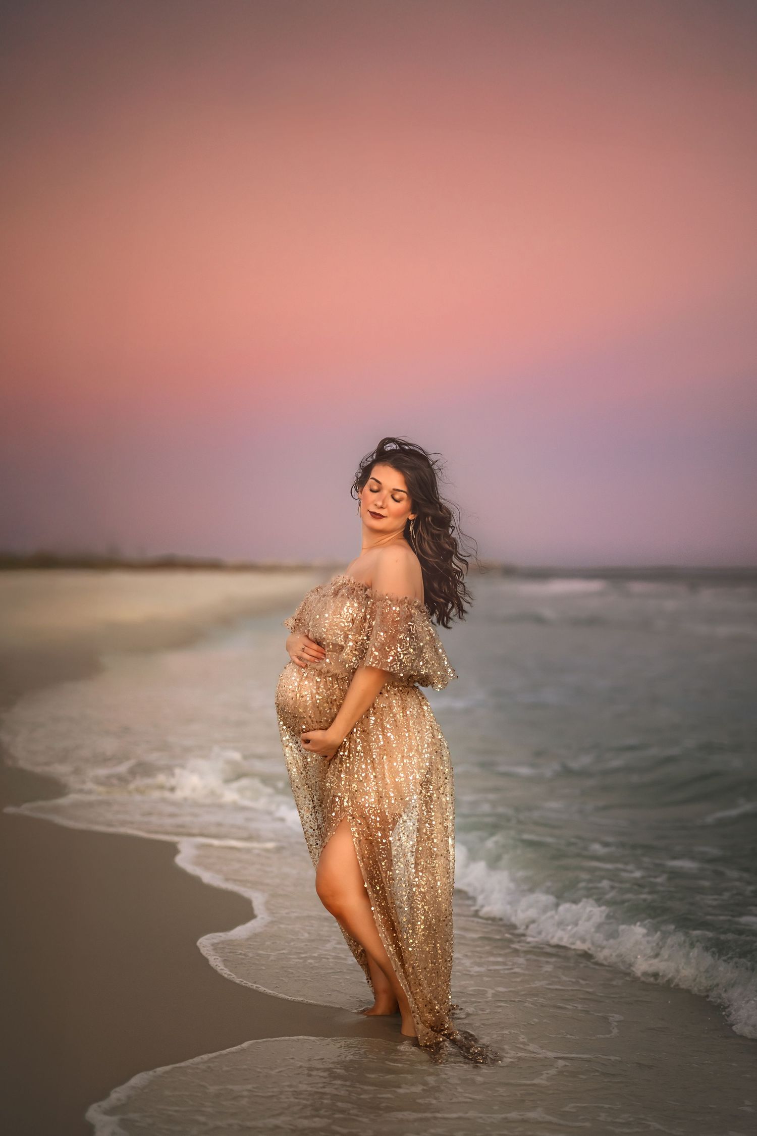 Pregnant woman in sparkly beige gown standing in ocean waves at sunset on a pink-hued beach.