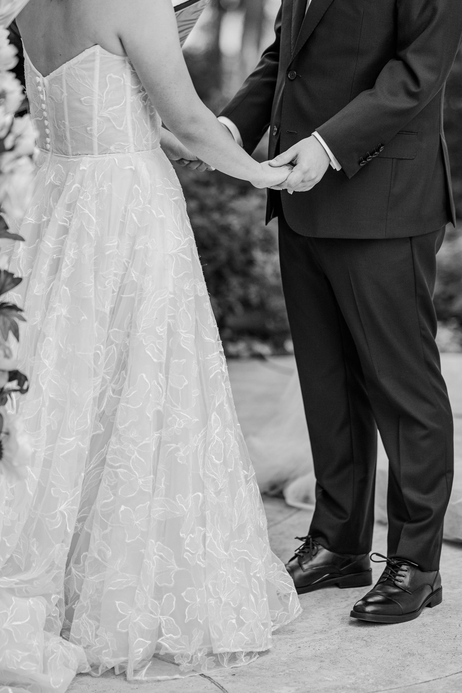 Black and white photo of bride and groom holding hands during wedding ceremony, bride in lace gown.