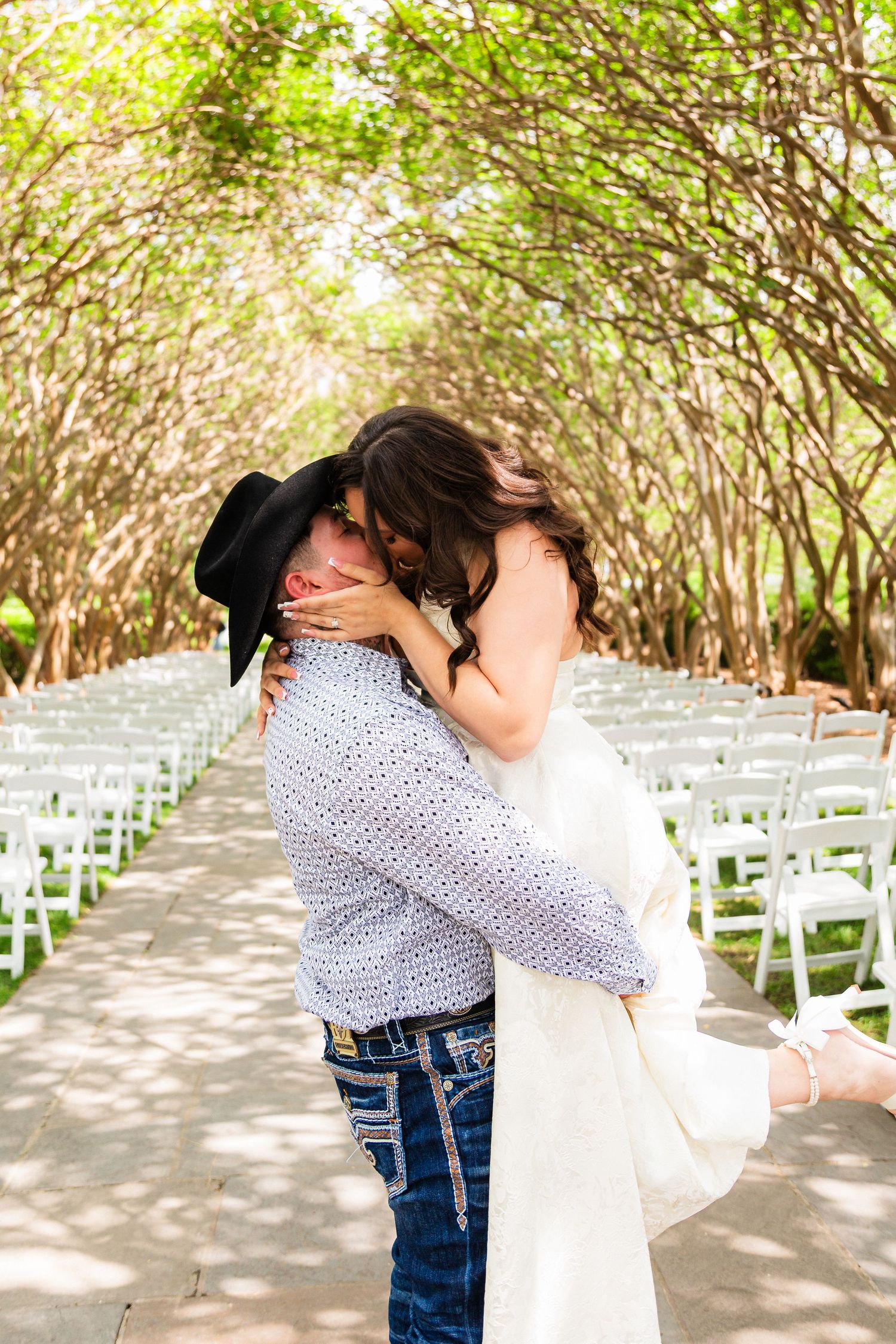 Groom in cowboy hat lifts bride in white dress, sharing a kiss along a tree-lined outdoor wedding ceremony aisle.