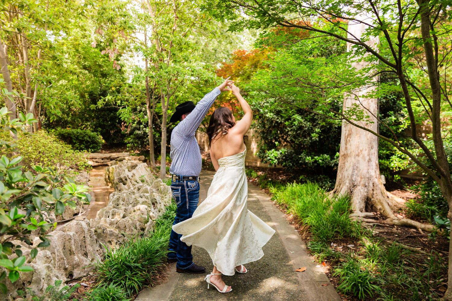 Couple dancing on a garden path surrounded by lush green trees, bride in flowing white gown being twirled by groom.