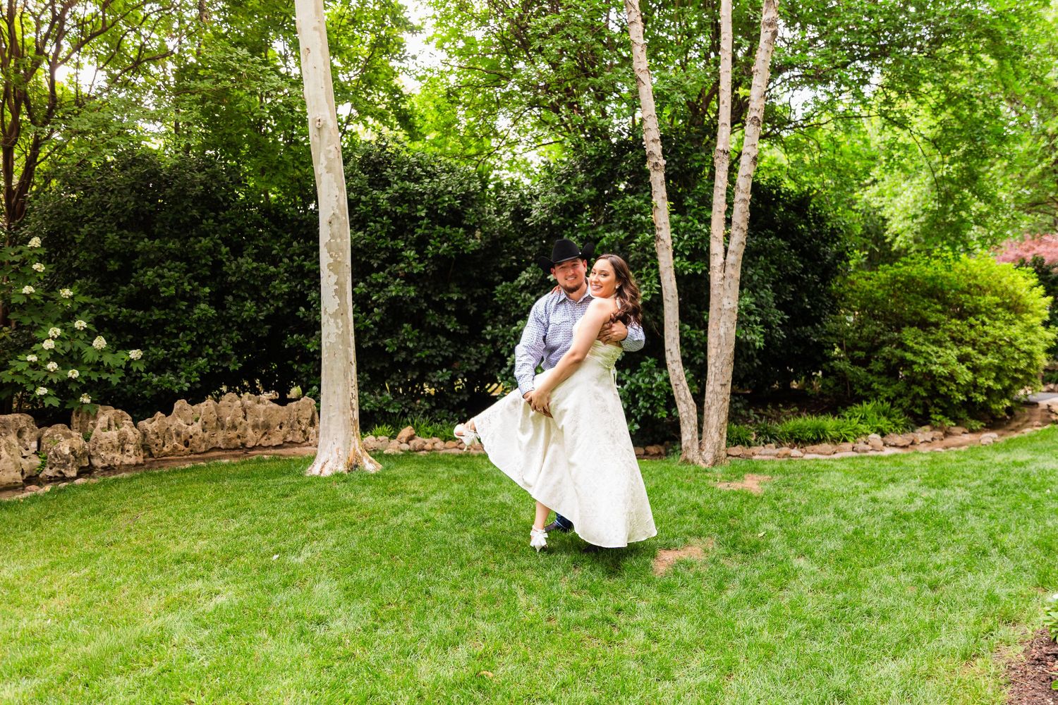 Groom lifting bride in white dress outdoors on lush green lawn surrounded by trees at wedding celebration.