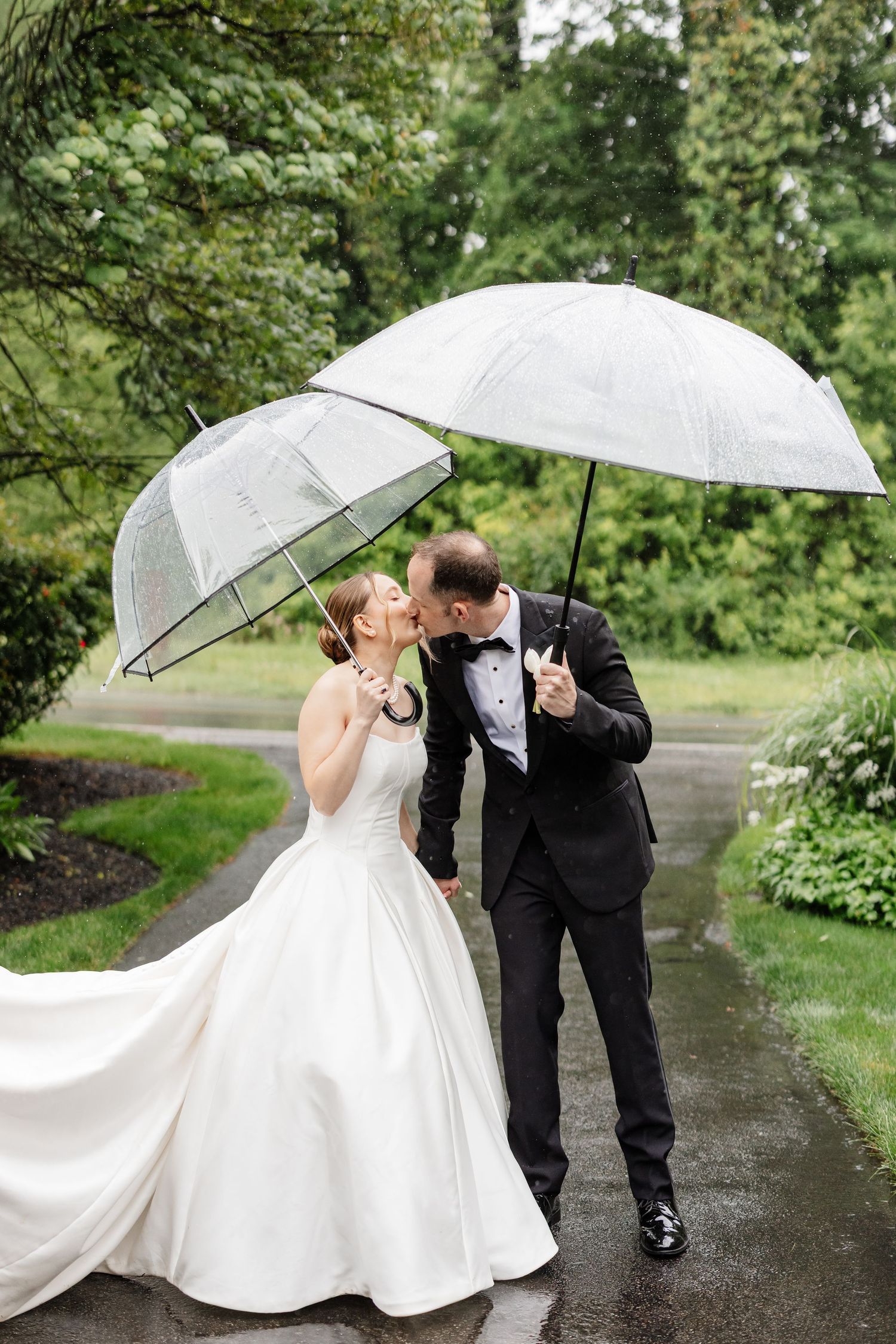 Bride and groom kiss while holding clear umbrellas on a rain-soaked path surrounded by lush green trees.