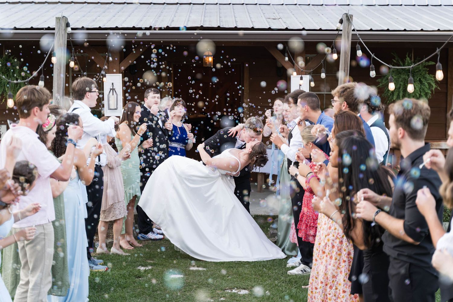 Groom dips bride during joyful outdoor wedding exit with guests blowing bubbles near rustic barn venue.