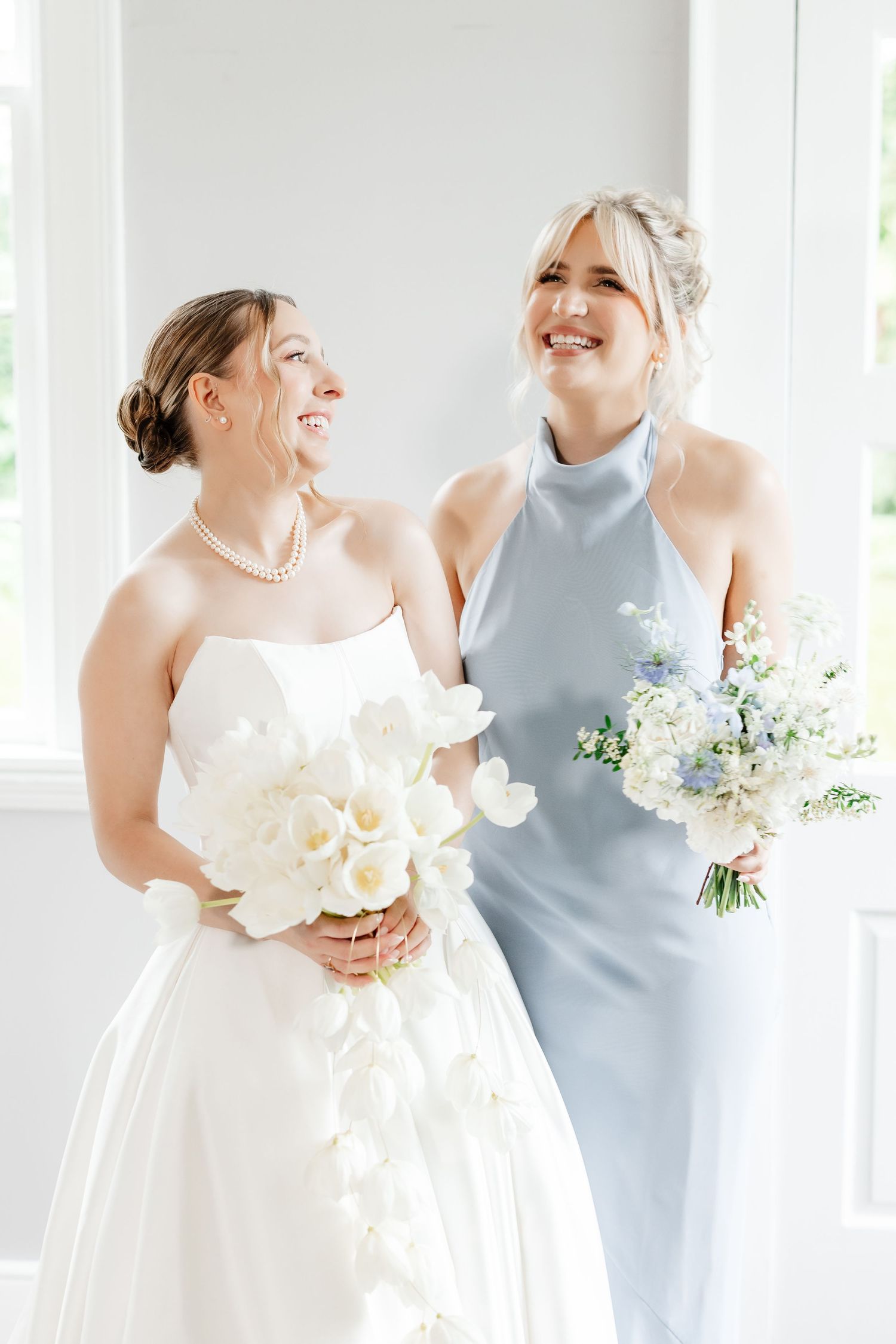 Bride in strapless white gown with pearl necklace and bridesmaid in blue halter dress holding floral bouquets.