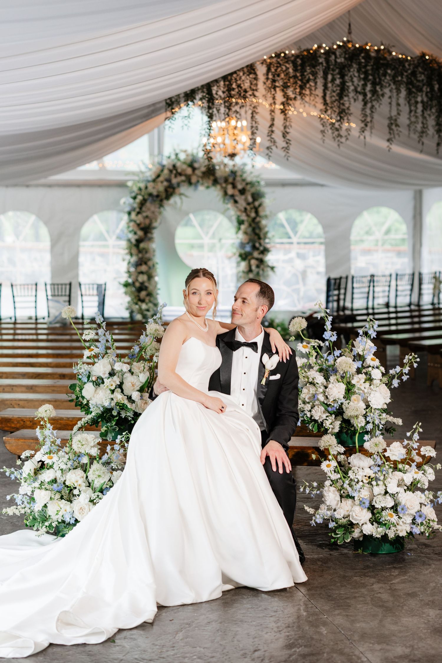 Bride and groom pose before a floral arch with white and blue flowers inside an elegant wedding tent.