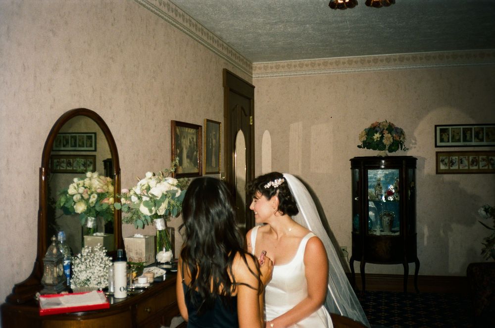 A bride in a white gown and veil shares a quiet moment with a guest inside a vintage room.