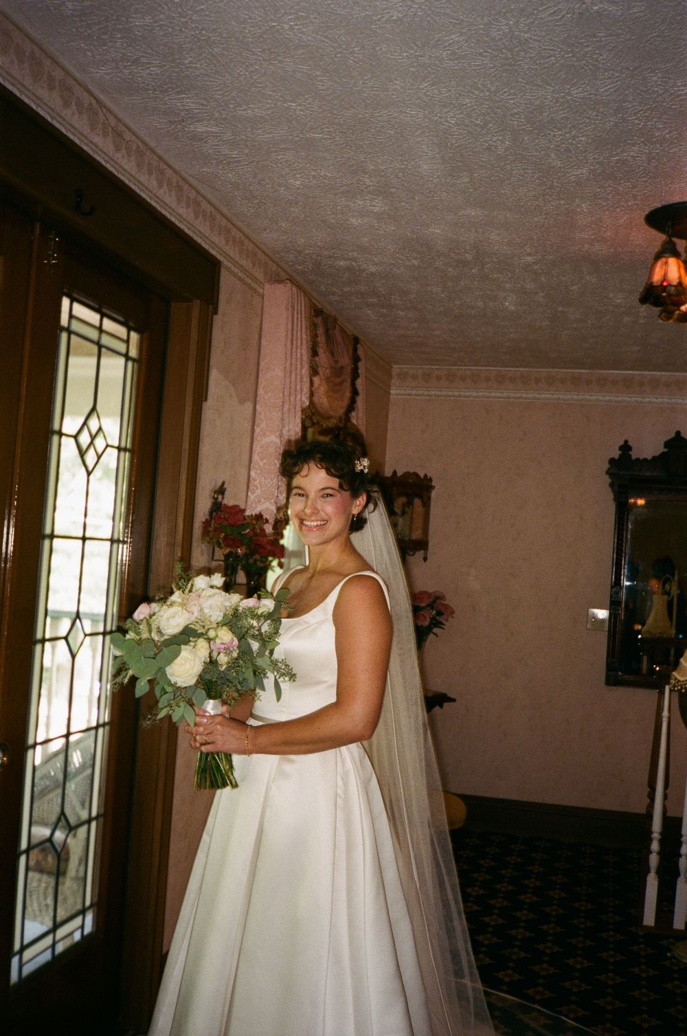A smiling bride in a white gown holds a bouquet of white roses inside a vintage home.