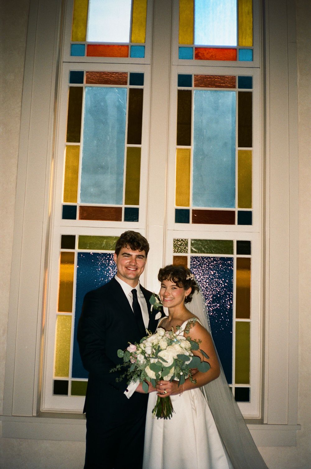 A bride and groom pose together in front of colorful stained glass windows on their wedding day.