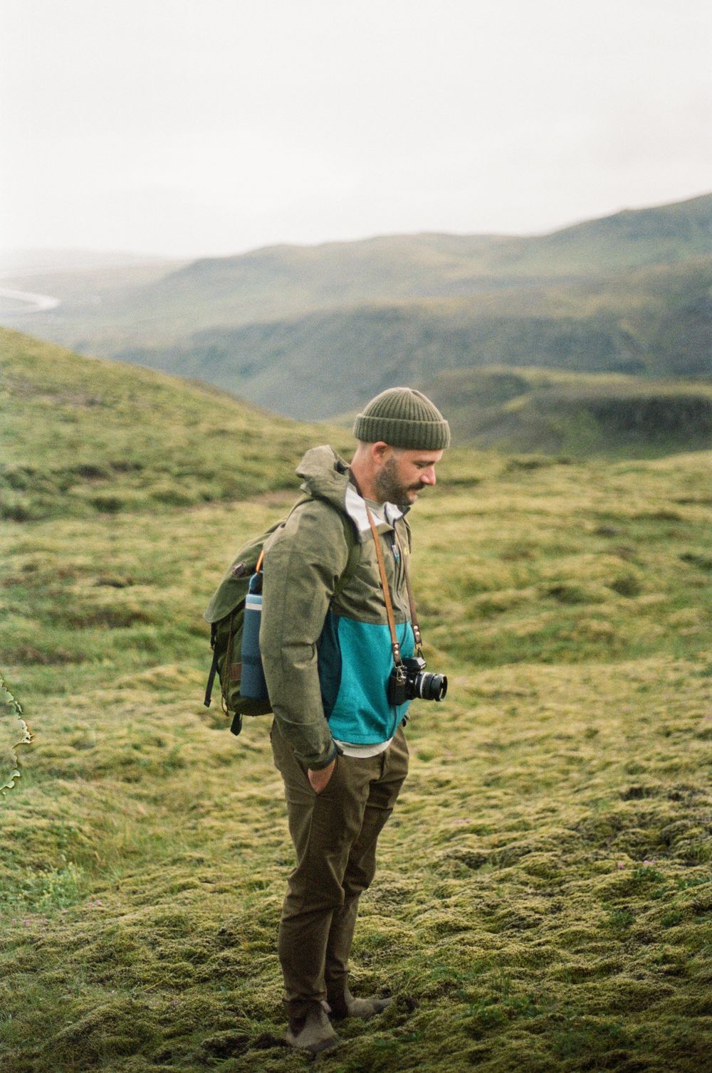 Male hiker with backpack and camera standing on green mossy hillside in misty mountain landscape.