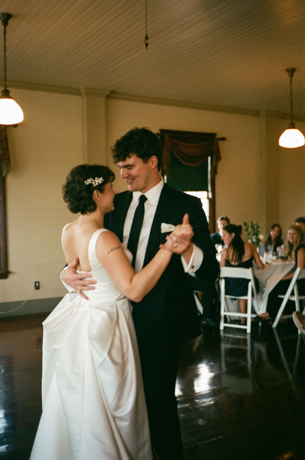 Bride and groom sharing their first dance in elegant ballroom with pendant lighting and guests seated.