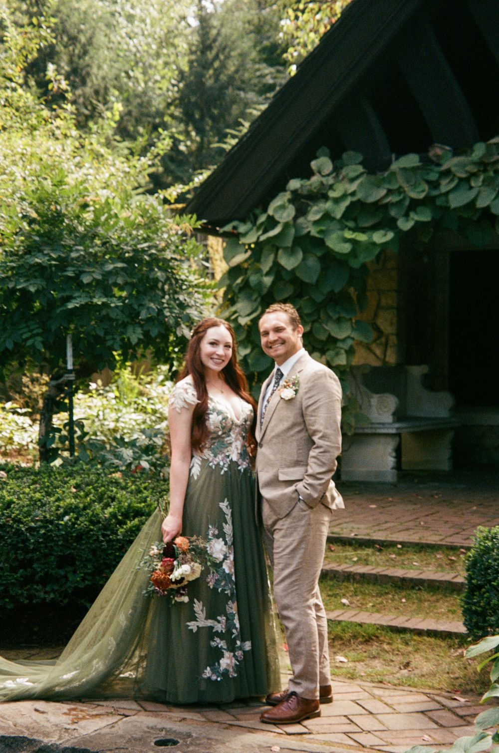 Bride in green floral gown and groom in tan suit posing in lush garden setting.