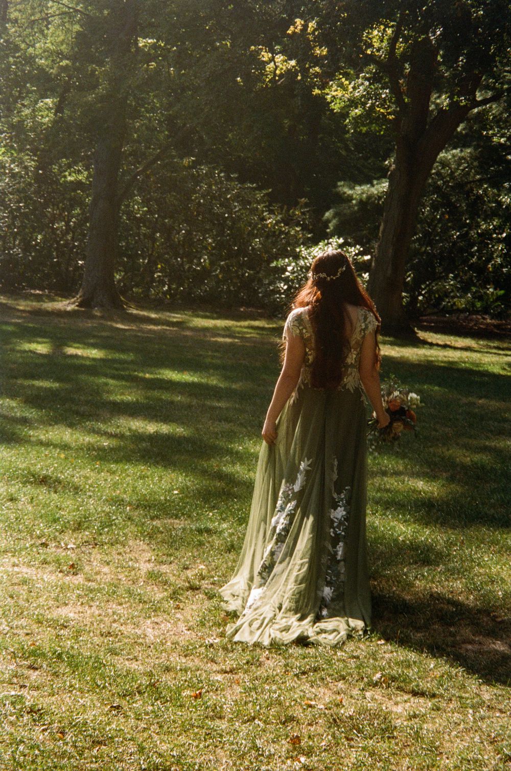Bride in flowing green embroidered gown with floral crown walking through sunlit garden forest.