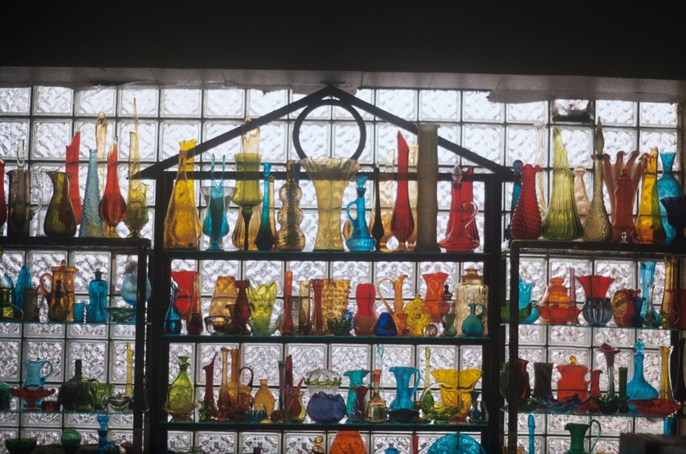 Colorful collection of vintage glass vases and bottles displayed on shelves in front of a window.